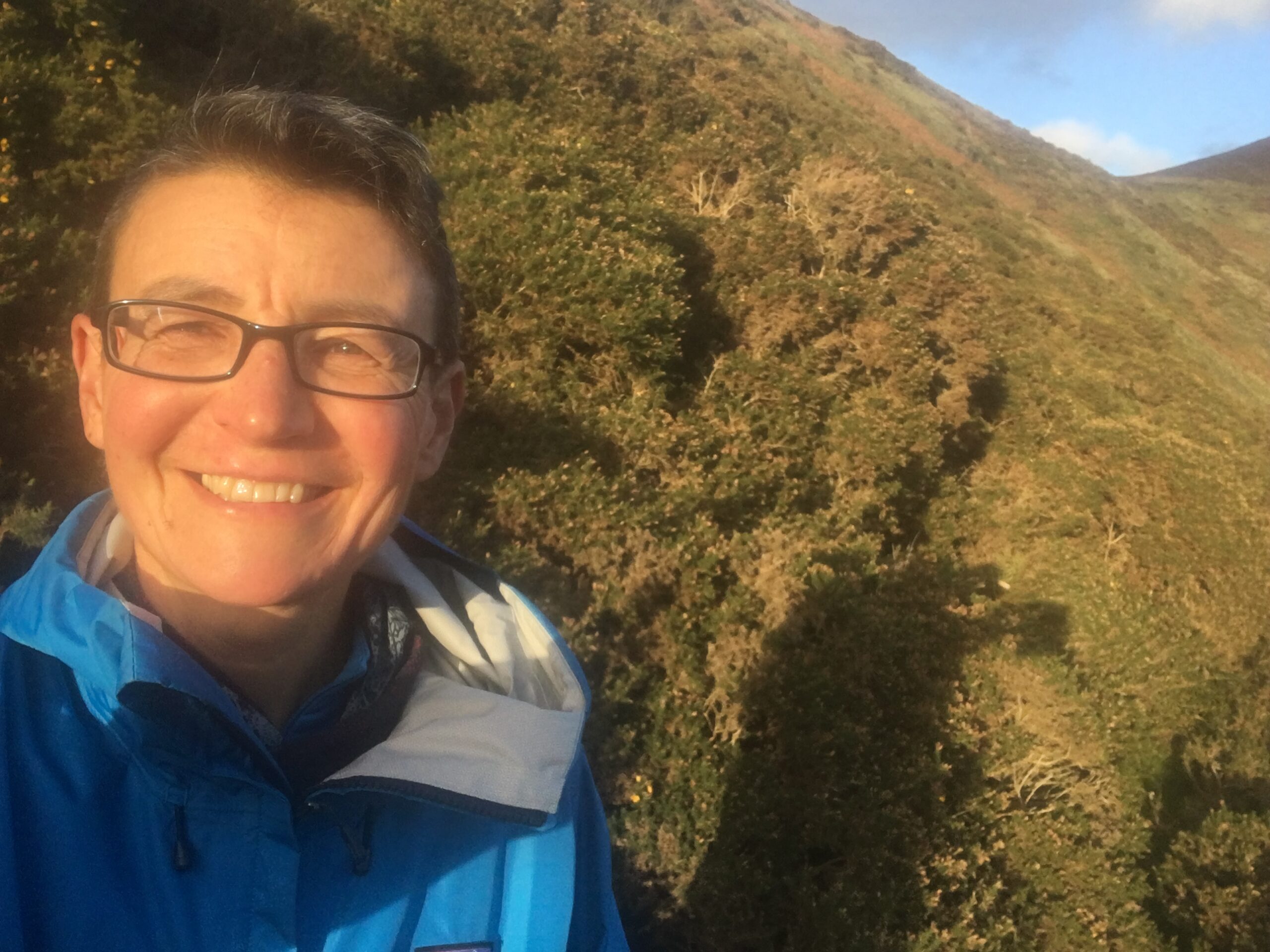 Lucy in blue raincoat with hills in the background, smiling and squinting.